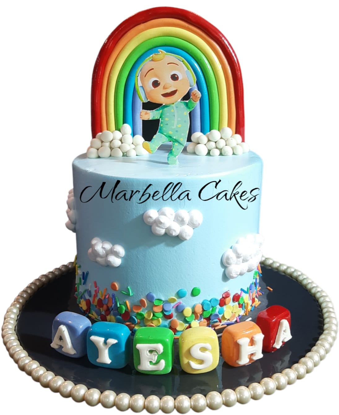 CoComelon Rainbow Sky Cake with blue buttercream, fondant rainbow topper, JJ character cutout, cloud decorations, and personalized name letter blocks