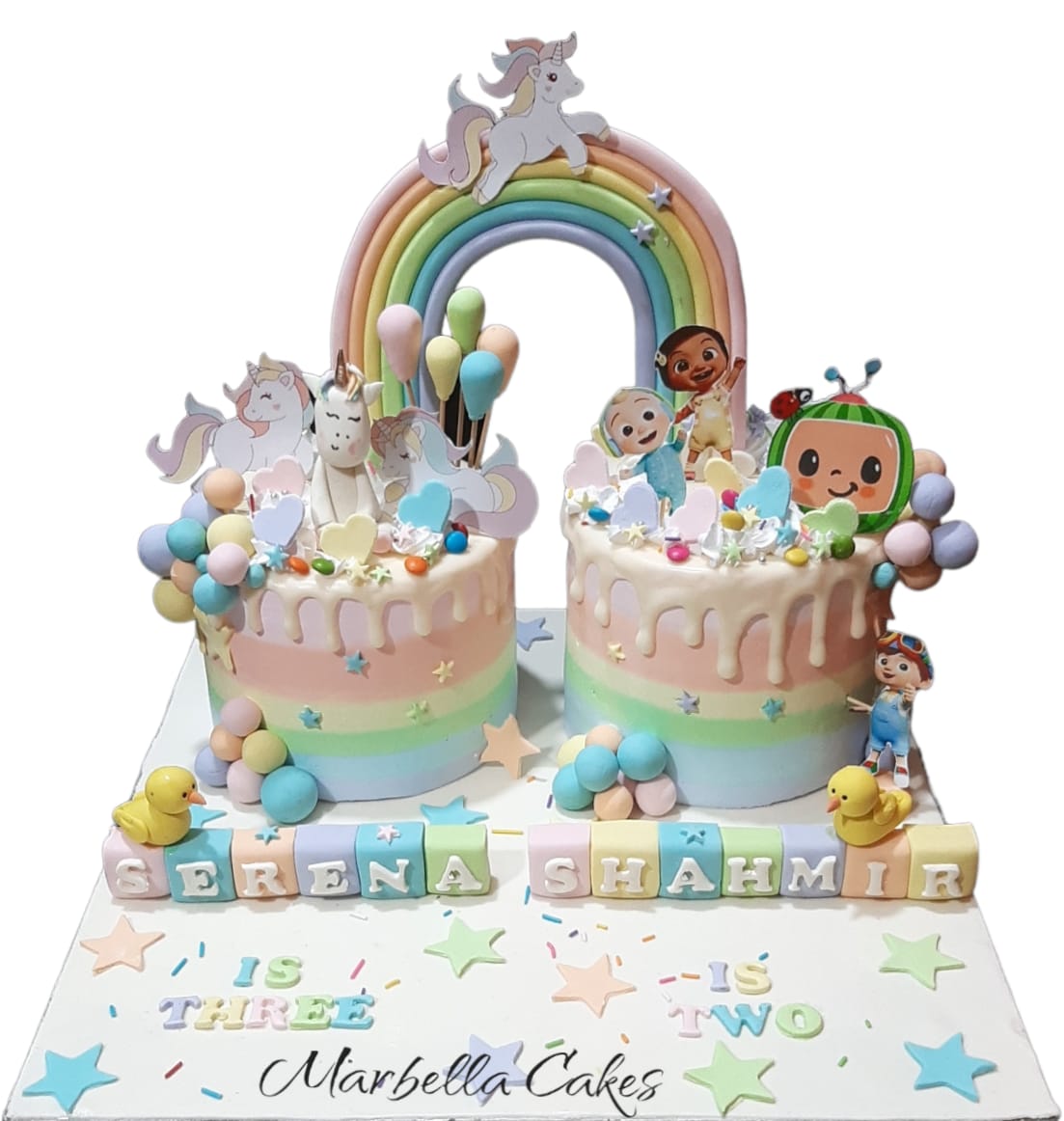 Twin birthday cake with unicorn theme on the left and CoComelon theme on the right, connected by a pastel rainbow arch, with personalized name blocks and age labels on the board