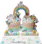 Twin birthday cake with unicorn theme on the left and CoComelon theme on the right, connected by a pastel rainbow arch, with personalized name blocks and age labels on the board