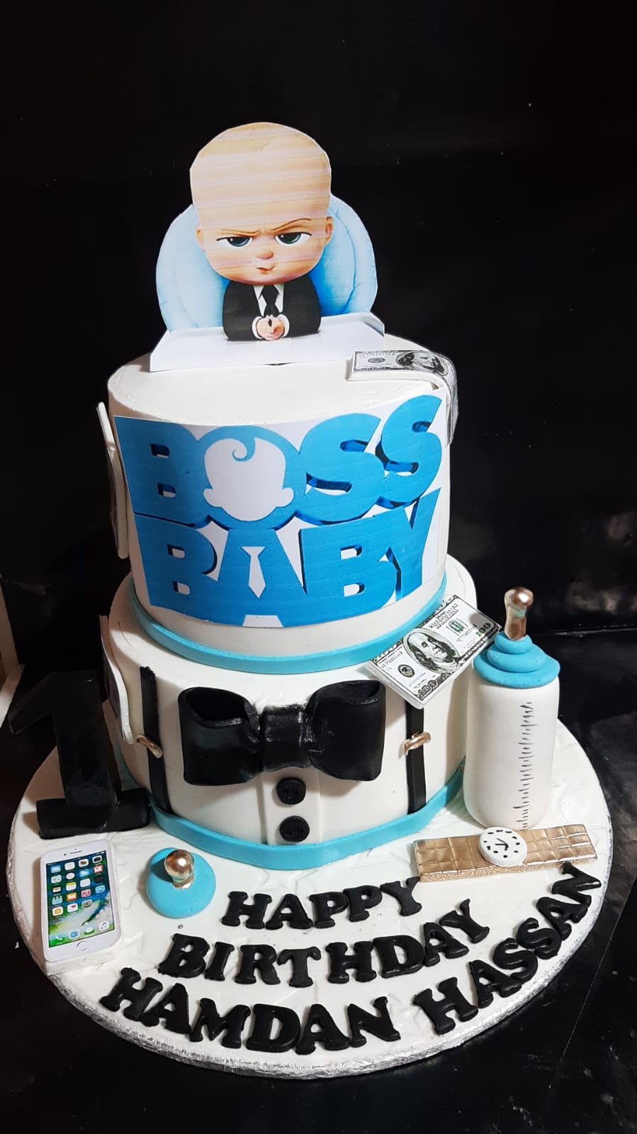 Boss Baby 2-tier birthday cake with white tuxedo bottom tier, BOSS BABY lettering on top tier, Boss Baby edible cutout topper, fondant dollar bills, baby bottle, smartphone and chocolate bar props