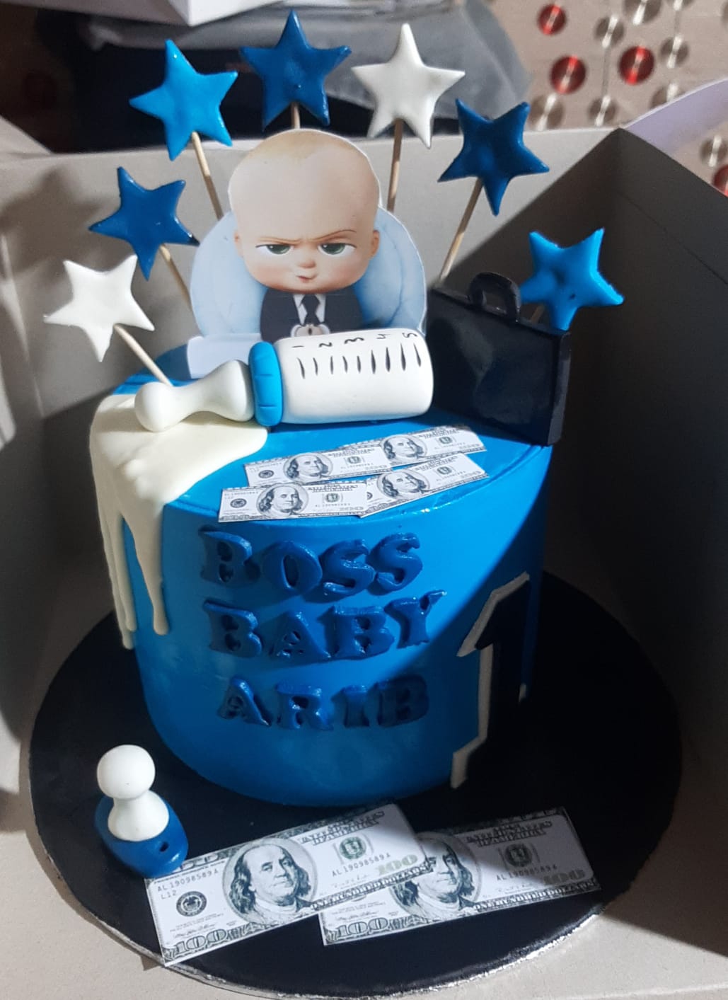 Boss Baby blue drip birthday cake with dollar money decorations, star toppers on sticks, and character topper