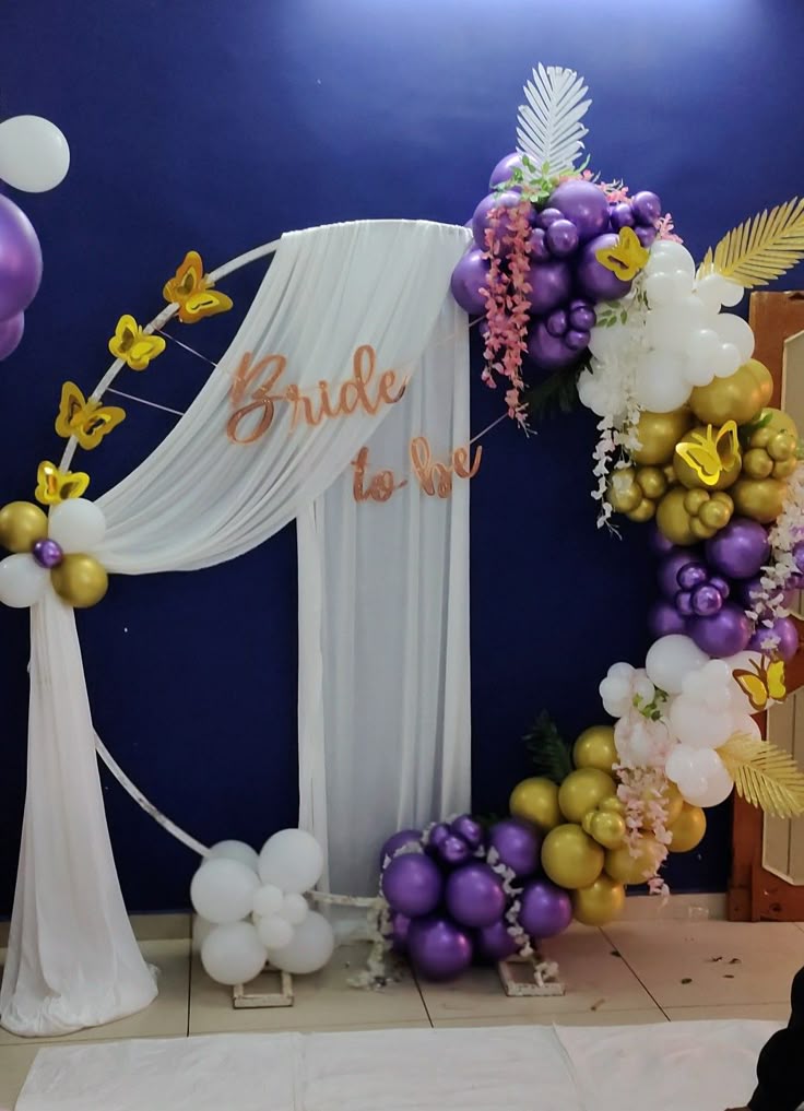 Bride to be bridal shower decoration with purple gold white balloon arch, butterfly cutouts and white sheer drape backdrop