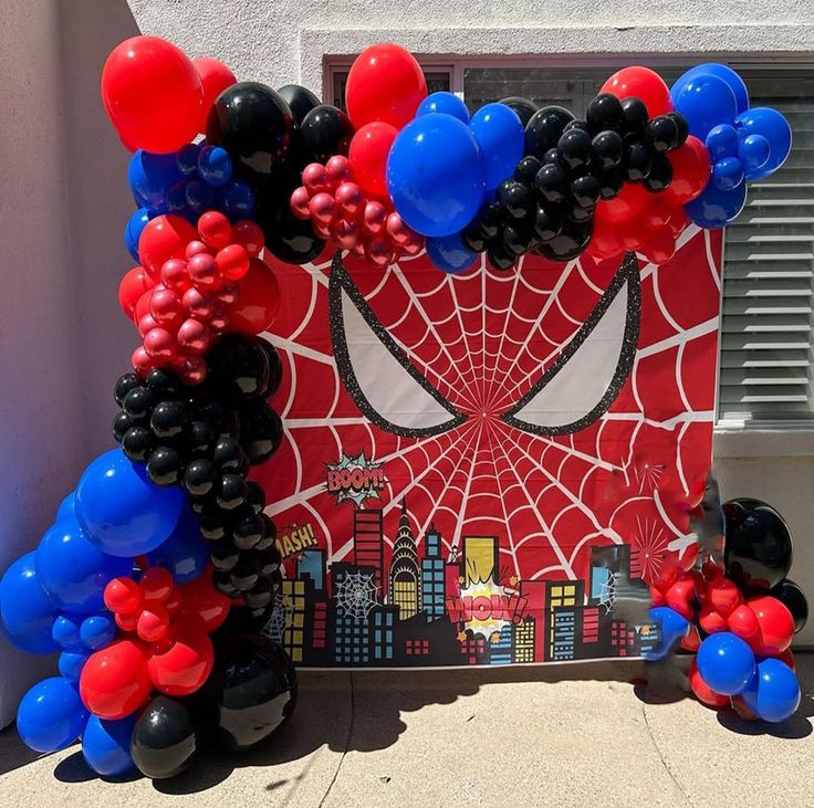 Spider-Man birthday decoration with red black blue balloon garland arch and Spider-Man spiderweb mask backdrop with city skyline and comic pop art accents