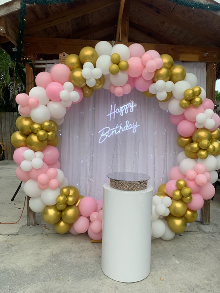 Pink white and gold balloon ring arch birthday decoration with Happy Birthday neon sign, white shimmer drape backdrop and white cylinder cake stand