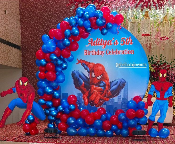 Spider-Man birthday decoration with large circular Spider-Man action backdrop, red and chrome blue balloon garland and two Spider-Man character standees on either side