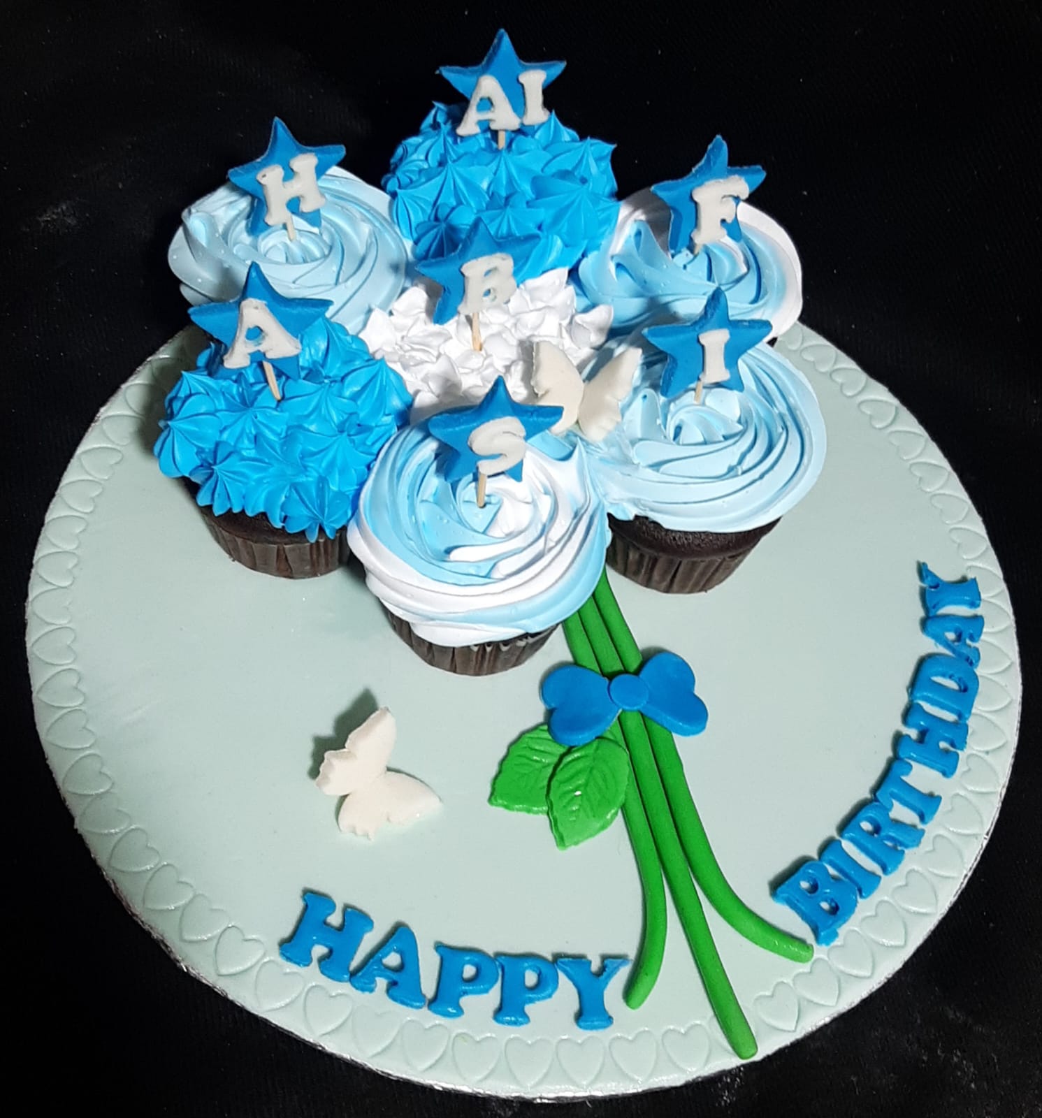 Happy Birthday pull-apart cupcake bouquet with blue and white swirl buttercream cupcakes, blue star fondant toppers with name letters arranged on a round mint fondant cake base with Happy Birthday lettering and green fondant flower stem
