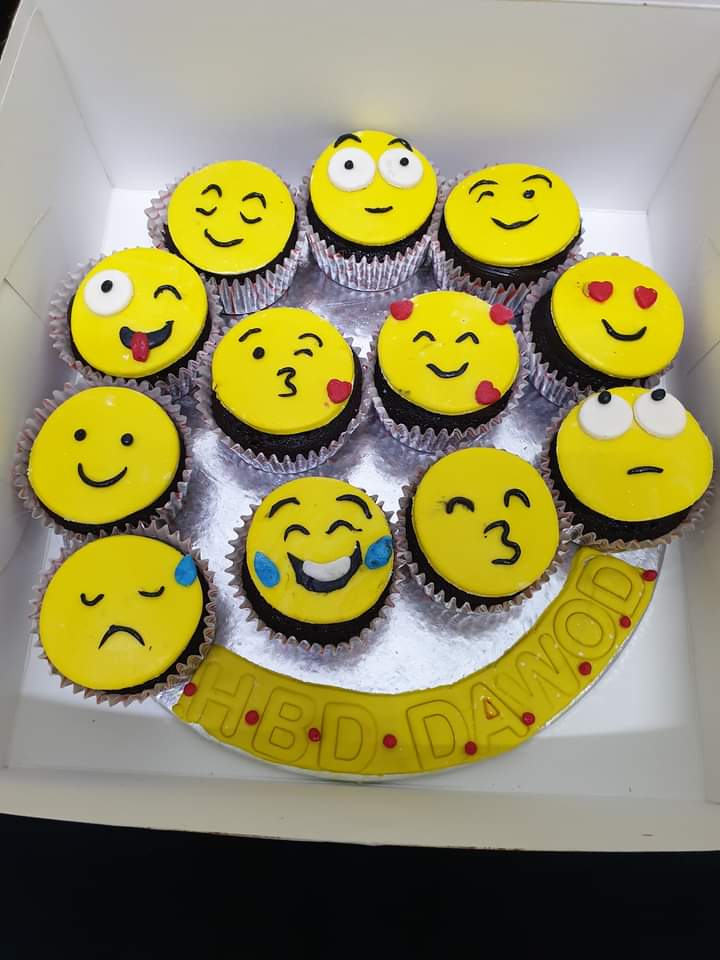 Box of 12 emoji birthday cupcakes with yellow fondant discs featuring 12 different hand-drawn emoji faces including happy sad laughing heart eyes and kissing, with personalized HBD name fondant banner at the bottom