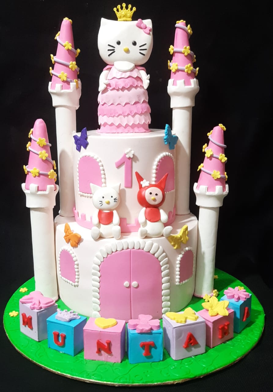 Hello Kitty princess castle 3-tier birthday cake in pink and white fondant with four castle towers, Hello Kitty crown topper, bunny figurines, pink arched door and personalized name alphabet blocks on green grass base