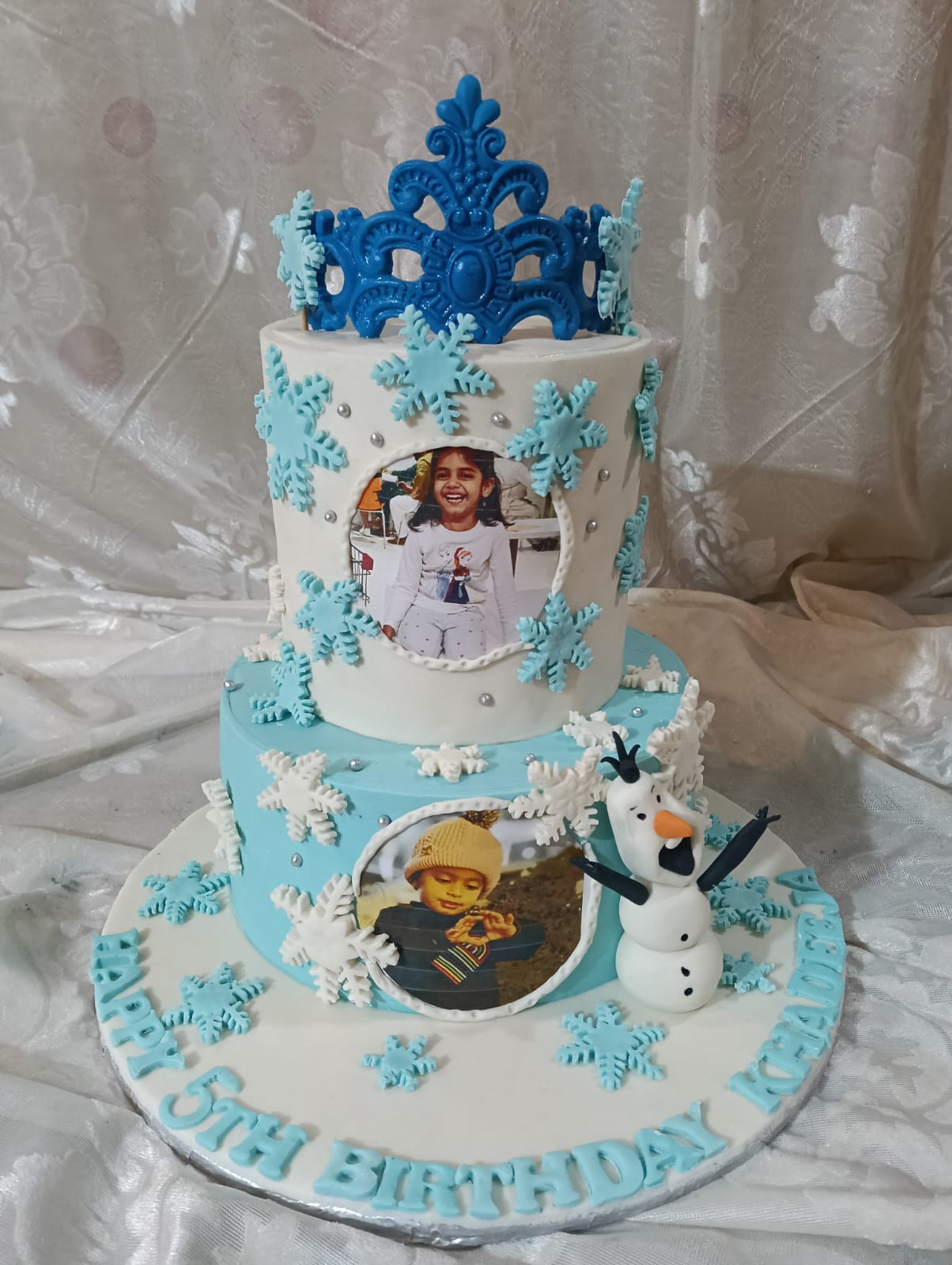 Frozen theme 2-tier birthday cake in blue and white fondant with snowflake decorations, Olaf snowman figurine, ornate blue crown topper, two round edible photo prints and personalized Happy Birthday name lettering on base