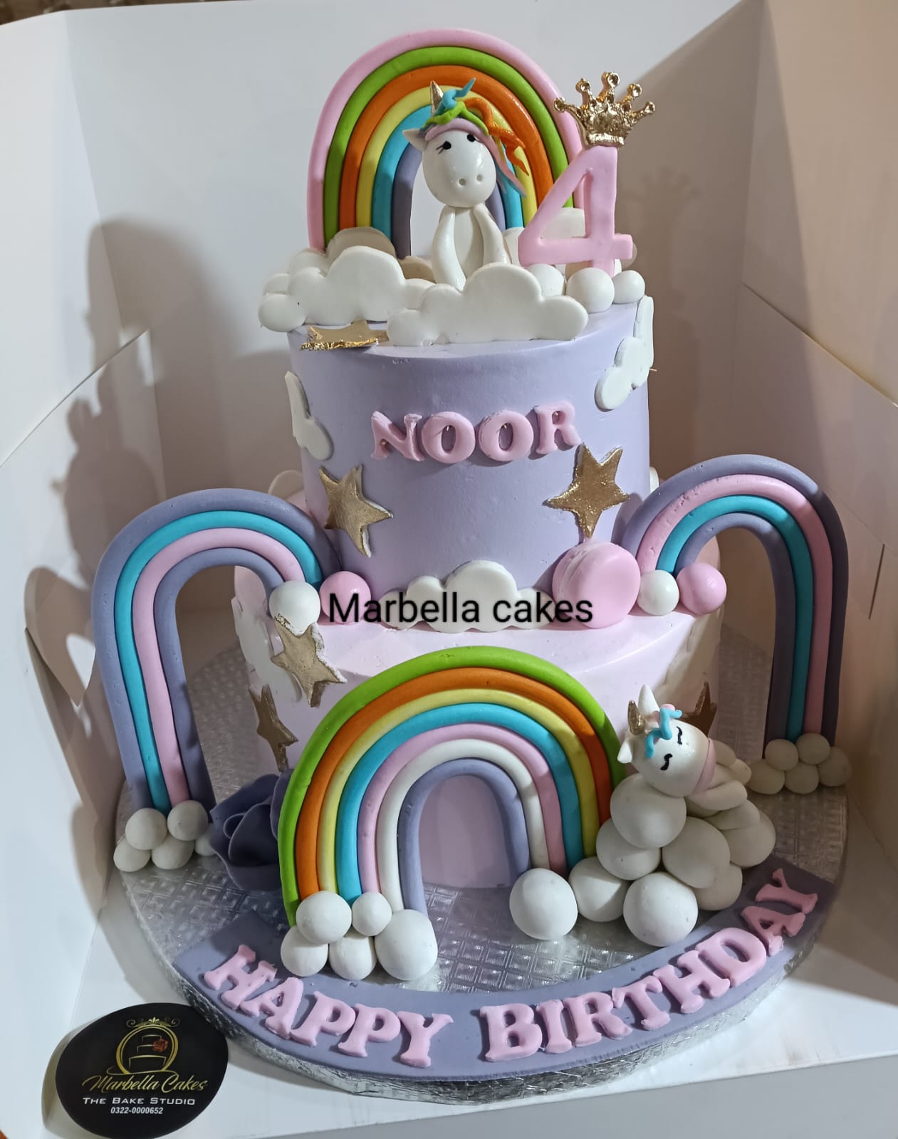 Unicorn and rainbow 2-tier lavender fondant birthday cake with pastel rainbow arches, white cloud ball border, cute unicorn figurines, gold star accents, personalized pink name lettering and Happy Birthday on base
