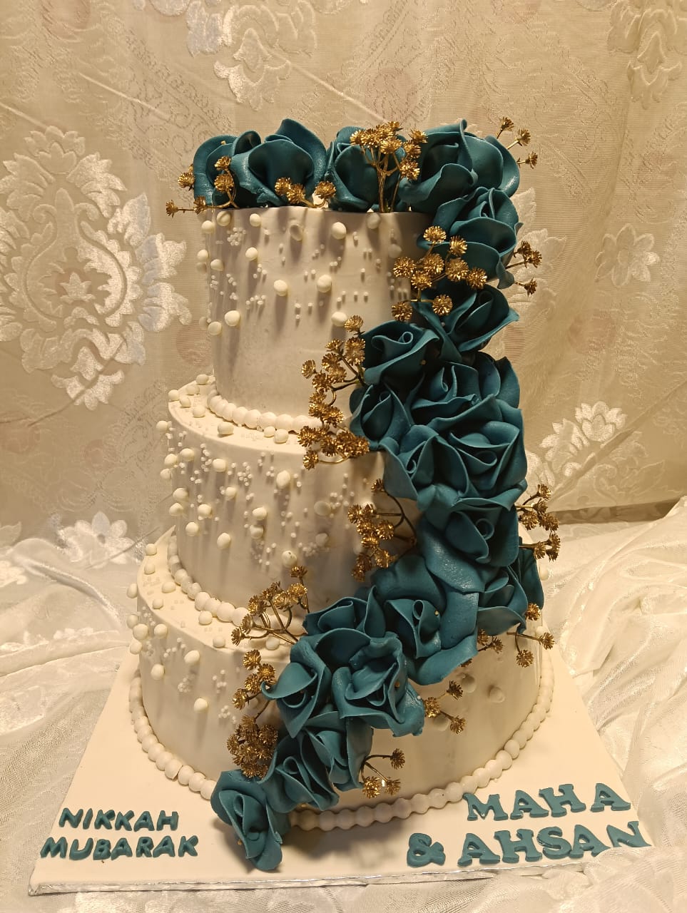Nikah Mubarak 3-tier white fondant wedding cake with cascading teal rose waterfall, gold gypsophila sprigs, white pearl dot trailing pattern, pearl bead borders and personalized Nikkah Mubarak couple names in teal lettering on base