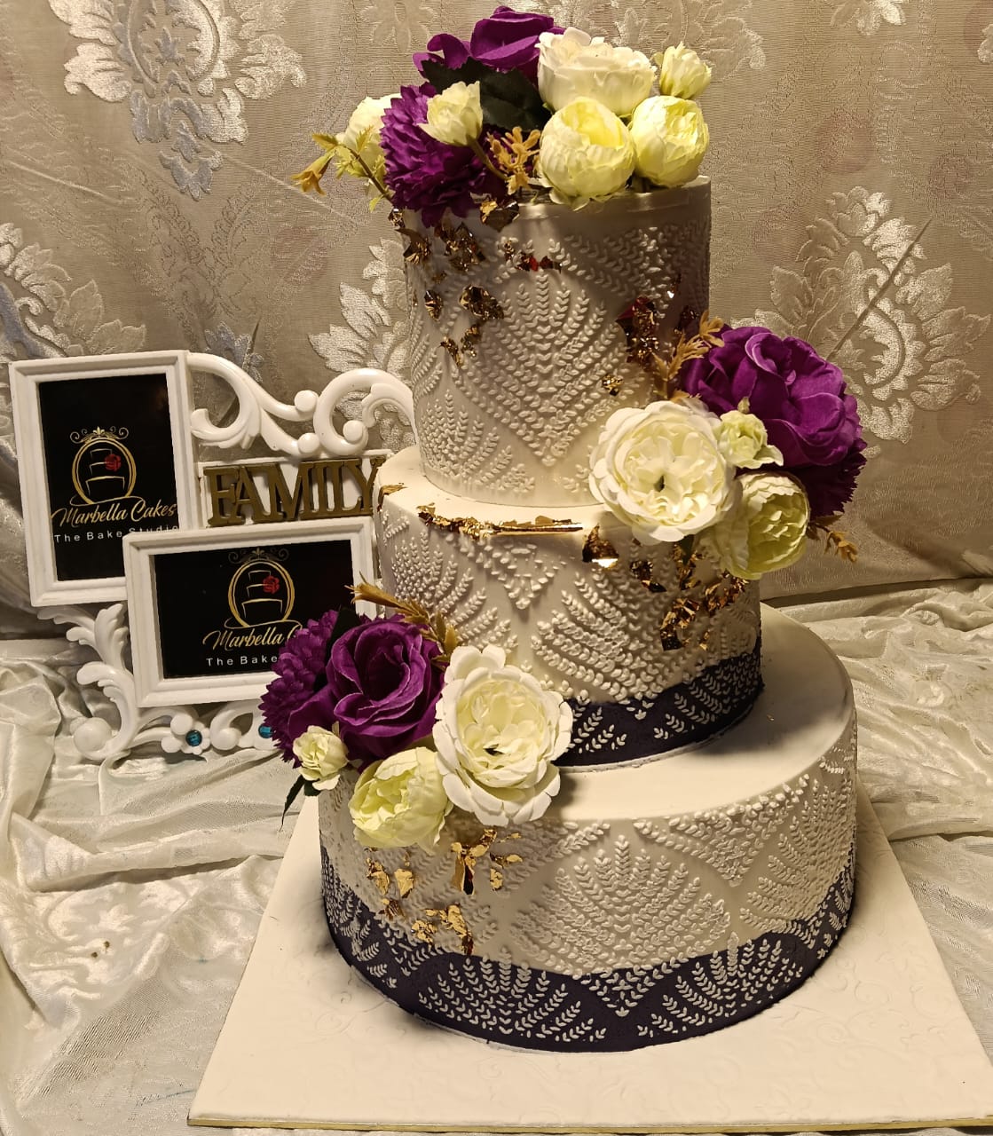Ivory and purple peony 3-tier wedding cake with white lace diamond and leaf embossed fondant, navy base borders, lush purple and cream peony clusters cascading down the tiers and gold leaf accents