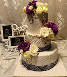Ivory and purple peony 3-tier wedding cake with white lace diamond and leaf embossed fondant, navy base borders, lush purple and cream peony clusters cascading down the tiers and gold leaf accents